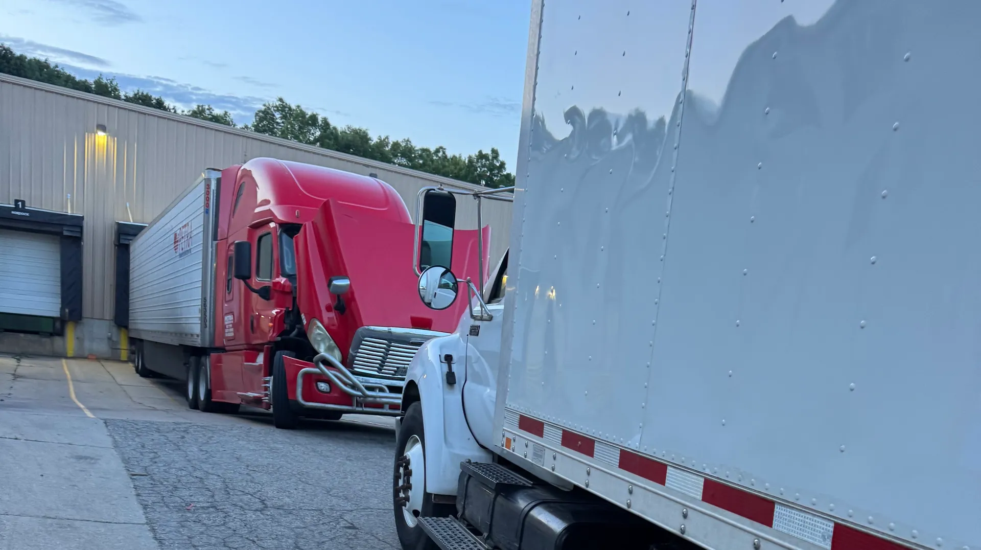 Red semi cab and white box truck parked at a warehouse with roll-up doors